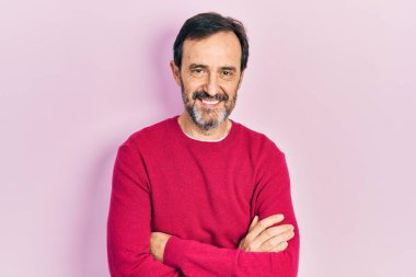 Orta yaşlı İspanyol bir adam sıradan kıyafetler giyip kollarını kavuşturup kameraya bakarken gülümsüyor. pozitif kişi. 