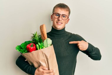 Young caucasian man holding paper bag with groceries pointing finger to one self smiling happy and proud 