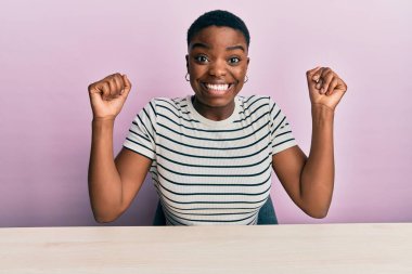 Young african american woman wearing casual clothes sitting on the table celebrating surprised and amazed for success with arms raised and open eyes. winner concept. 