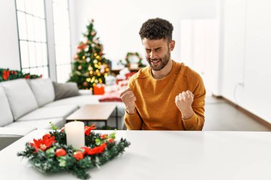 Arab young man sitting on the table by christmas tree very happy and excited doing winner gesture with arms raised, smiling and screaming for success. celebration concept. 