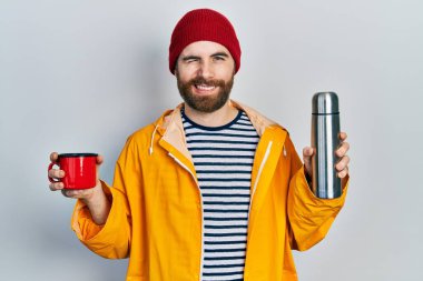 Sakallı, elinde kahve fincanı olan beyaz bir adam kameraya seksi bir ifadeyle bakıyor. Neşeli ve mutlu bir yüzü var.. 