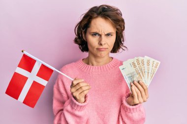 Elinde Norveç bayrağı ve Krone banknotları tutan genç esmer kadın şüpheci ve gergin, sorun yüzünden surat asıyor. negatif kişi. 