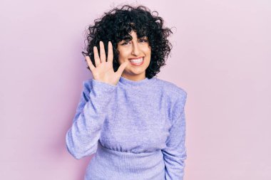 Orta Doğulu genç bir kadın gündelik kıyafetler giyer ve beş numarayı göstererek gülümserken kendinden emin ve mutlu görünürdü.. 