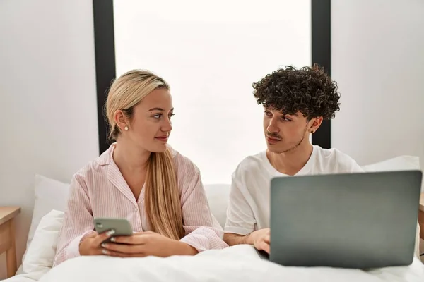 Młoda Piękna Para Pomocą Laptopa Smartfona Siedzi Łóżku Domu — Zdjęcie stockowe