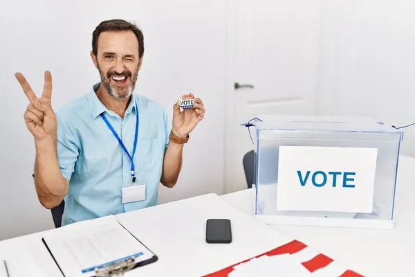 私は指で勝利のサインを行うカメラでの幸せな顔のウィンクで笑顔投票バッジを保持投票によって座ってひげを持つ中年の男 — ストック写真