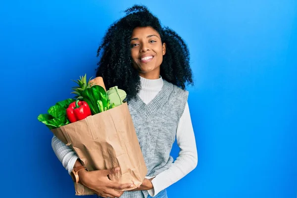 アフリカ系アメリカ人の女性アフロヘア保持紙袋で食料品と肯定的かつ幸せな立っていると歯を示す自信を持って笑顔で笑顔 — ストック写真