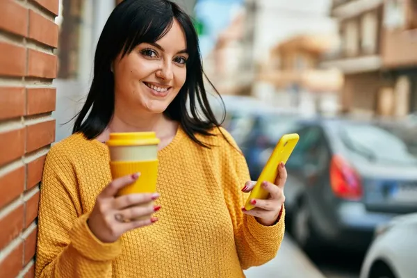 年轻的黑发女人高兴地笑着 一边用智能手机 一边靠在砖墙上喝咖啡 — 图库照片