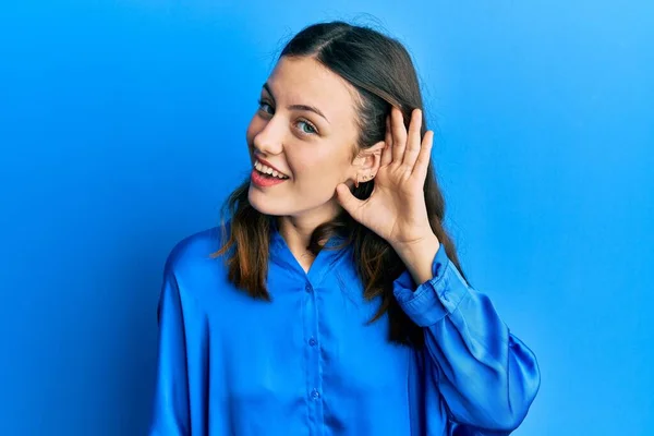 若いブルネットの女性は噂やゴシップに耳を傾ける耳の上に手で笑みを浮かべてカジュアルな青いシャツを着ている 聴覚障害の概念 — ストック写真