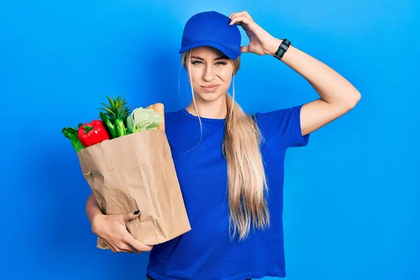 年轻的高加索女人穿着信使制服 带着超市的食品杂货 对问题感到困惑和疑惑 拿不定主意 手拿着头思考 沉思的概念 — 图库照片