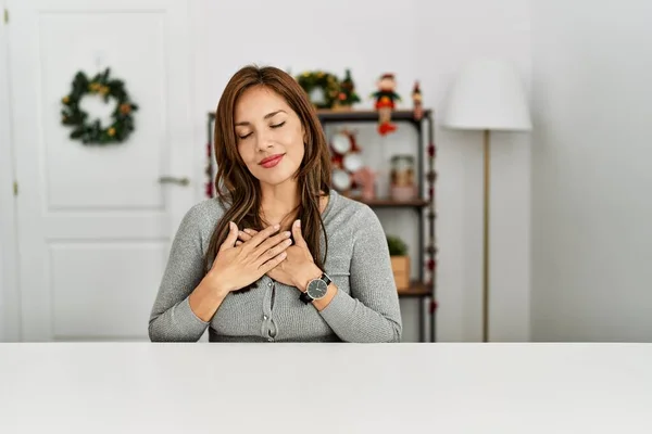年轻的拉丁女人坐在桌上 戴着圣诞装饰品 双手放在胸前笑着 闭着眼睛 脸上挂着感激的手势 健康概念 — 图库照片