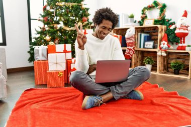 Genç Afro-Amerikan adam dizüstü bilgisayar kullanıyor Noel ağacının yanında oturmuş kameraya gülümsüyor ve zafer işareti yapıyor. İki numara.. 