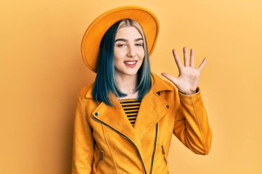 Sarı şapkalı ve deri ceketli genç modern bir kız beş numaralı parmaklarıyla onu gösterirken kendinden emin ve mutlu bir şekilde gülümsüyor.. 