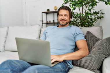 Orta yaşlı, beyaz bir adam evinde dizüstü bilgisayar kullanarak gülümsüyor.