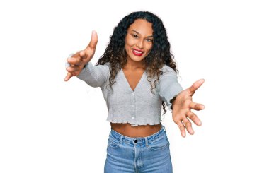 Kıvırcık saçlı, sıradan kıyafetler giyen genç İspanyol kadın sarılmak için kollarını açmış kameraya bakıyor. Mutluluğu kucaklayan neşeli bir ifade.. 