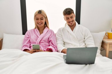 Genç beyaz çift mutlu bir şekilde dizüstü bilgisayar ve akıllı telefon kullanarak evde yatakta oturuyor..