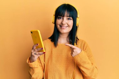 Genç İspanyol kadın akıllı telefon kullanıyor kulaklık takıyor gülümsüyor mutlu bir el ve parmak ile işaret ediyor 