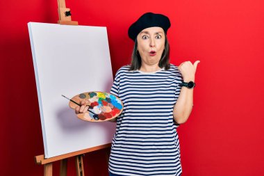 Orta yaşlı İspanyol kadın elinde paletle ayakta dikiliyor resim sehpası şaşırmış bir şekilde el ile işaret ediyor, ağzı açık şaşkın bir şekilde.. 