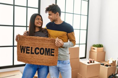 Genç Latin çift mutlu bir şekilde gülümsüyor. Yeni evimize hoş geldin paspası koyuyorlar..