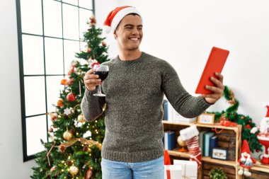 Young hispanic man make selfie by the touchpad drinking wine standing by christmas tree at home