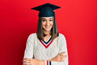 Mezuniyet şapkası takan, kollarını kavuşturup kameraya gülümseyen esmer kız. pozitif kişi. 