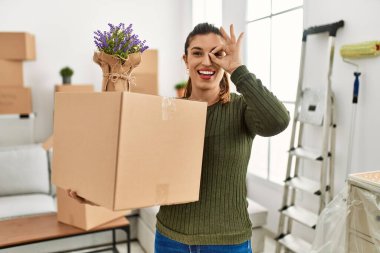 Young hispanic woman holding cardboard box at moving smiling happy doing ok sign with hand on eye looking through fingers 