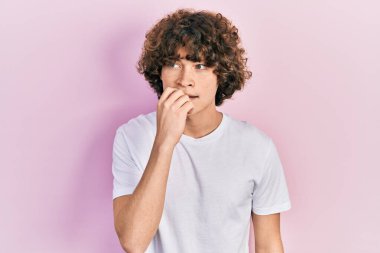 Handsome young man wearing casual white t shirt looking stressed and nervous with hands on mouth biting nails. anxiety problem. 