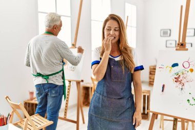 Hispanic woman wearing apron at art studio looking stressed and nervous with hands on mouth biting nails. anxiety problem. 