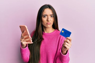 Genç İspanyol kız elinde akıllı telefon ve kredi kartı ile şüpheci ve gergin, sorun yüzünden surat asıyor. negatif kişi. 