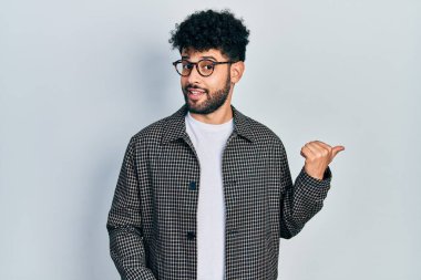 Young arab man with beard wearing glasses smiling with happy face looking and pointing to the side with thumb up. 