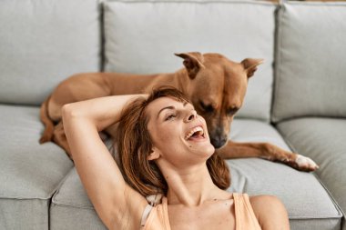 Beyaz bir kadın köpeğe sarılıyor. Evde yerde oturuyor.