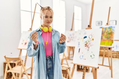 Young caucasian girl at art studio doing money gesture with hands, asking for salary payment, millionaire business 