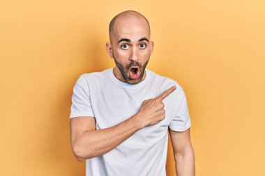 Gündelik beyaz t-shirt giyen genç kel adam parmaklarını yana doğru göstererek şaşırdı, ağzı açık hayret içinde kaldı.. 