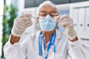 Doktor üniforması giyen yaşlı gri saçlı kadın ve klinikte aşıyı elinde tutan tıbbi maske.