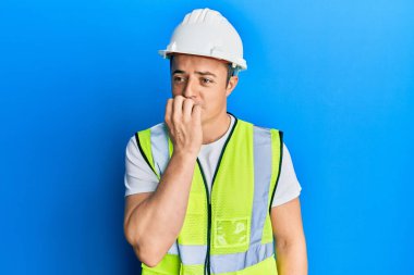 Handsome young man wearing safety helmet and reflective jacket looking stressed and nervous with hands on mouth biting nails. anxiety problem. 