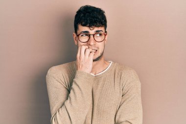 Günlük kıyafetler ve gözlük takan genç İspanyol adam gergin ve gergin görünüyor. Elleri ağızda, tırnaklarını yiyor. anksiyete. 