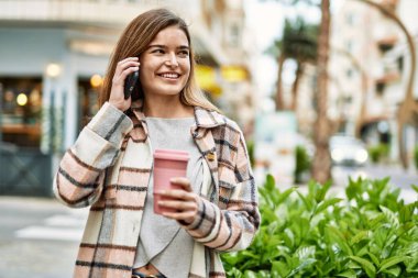 Elinde kahve tutan genç sarışın kadın caddede akıllı telefondan konuşuyor.