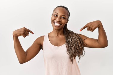 Saçları örgülü siyah bir kadın izole bir arka planda duruyor. Yüzünde bir gülümsemeyle, gururlu ve mutlu bir şekilde kendini işaret ediyor.. 