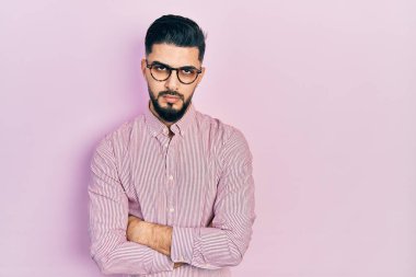 Sakallı, rahat gömlekli, gözlüklü, şüpheci ve gergin, yüzünde çapraz kollu tasvip etmeyen bir ifade olan yakışıklı bir adam. negatif kişi. 