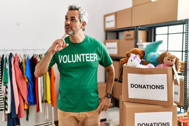 Middle age hispanic man wearing volunteer t shirt at donations stand thinking concentrated about doubt with finger on chin and looking up wondering 