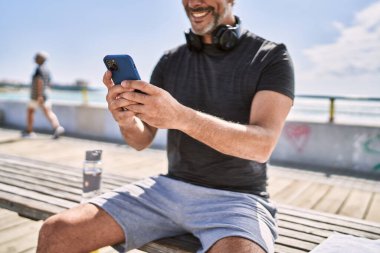 Orta yaşlı İspanyol adam deniz kenarında akıllı telefon kullanarak spor giyer.