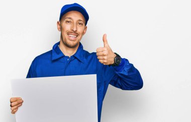 Bald man with beard wearing builder jumpsuit uniform holding empty banner smiling happy and positive, thumb up doing excellent and approval sign 
