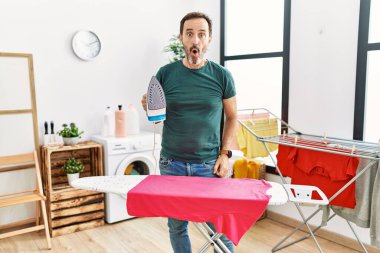 Orta yaşlı, sakallı, evde giysilerini ütüleyen ağzı açık, korkmuş ve şaşırmış bir adam. 