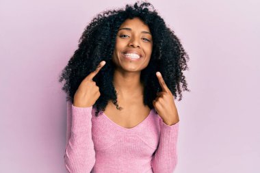 Afro saçlı, günlük pembe tişörtlü neşeli gülümseyen ve parmaklarıyla ve ağzıyla işaret eden Afro-Amerikalı kadın. diş sağlığı kavramı. 