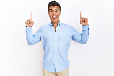 Young hispanic man wearing business shirt standing over isolated background smiling amazed and surprised and pointing up with fingers and raised arms. 