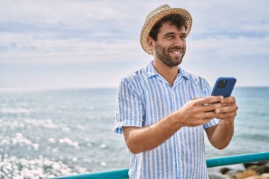 Genç İspanyol adam sahilde akıllı telefon kullanarak mutlu bir şekilde gülümsüyor.