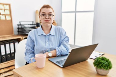 Ofiste çalışan genç kızıl saçlı kadın komik suratlı, dizüstü bilgisayarlı, şişkin yanaklar kullanıyor. Havayla şişmiş ağız, çılgın bir ifade.. 