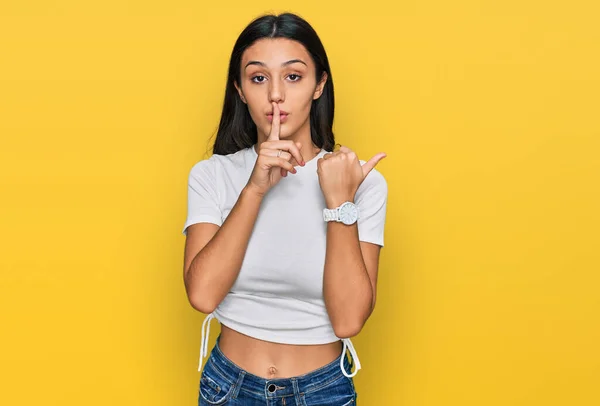 若いヒスパニック系の女の子の横に手を指して唇に指で静かにするように求めてカジュアルな白いTシャツを着て 沈黙と秘密の概念 — ストック写真