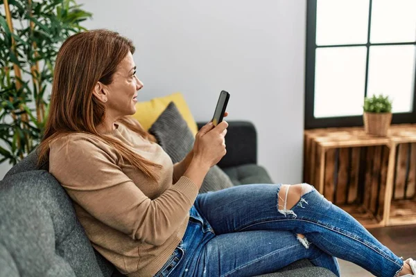 中年白人女性を使用してスマートフォンを使用してソファの上に家に座って — ストック写真