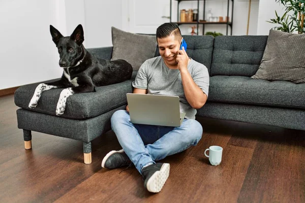 家で犬と一緒に床に座っているスマートフォンにノートパソコンを使用して若いヒスパニック系の男 — ストック写真