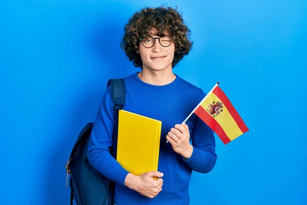 Handsome young man exchange student holding spanish flag winking looking at the camera with sexy expression, cheerful and happy face.